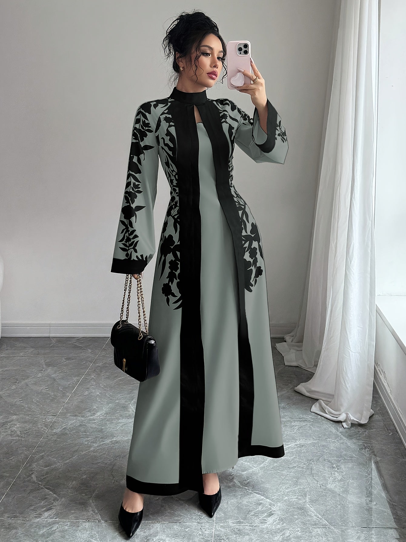 Woman taking a mirror selfie wearing a long, patterned dress in a room with light gray walls and white curtains.