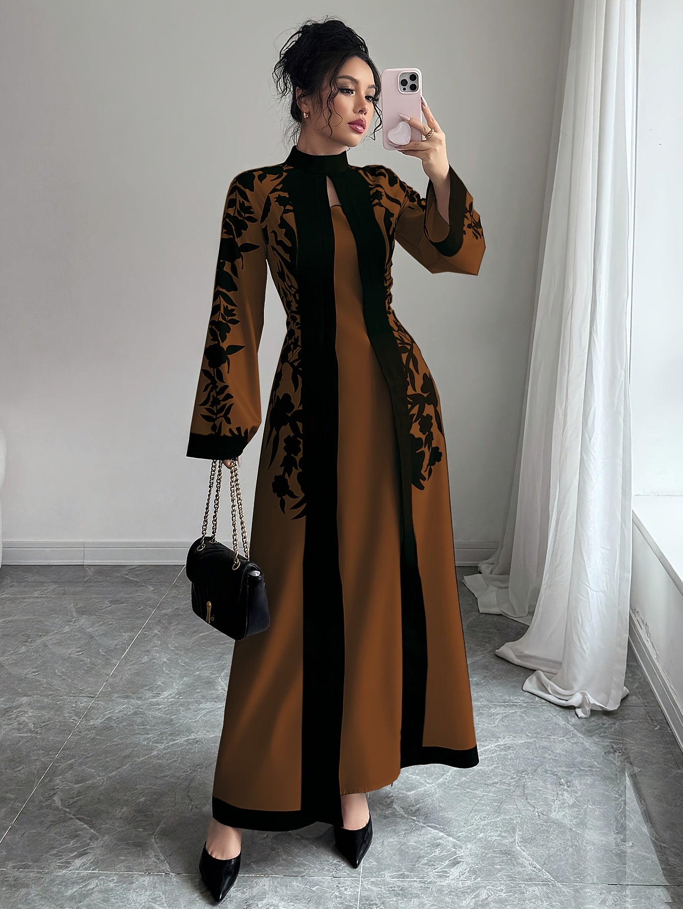 Woman taking a mirror selfie wearing a long brown and black dress in a room with light gray walls and white curtains.