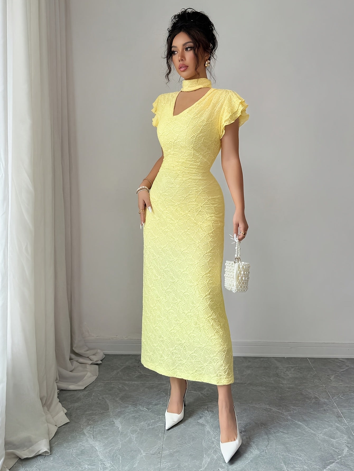 A woman models a pale yellow textured midi dress with a high mock neck, ruffled cap sleeves, and a keyhole cutout at the collar. She is wearing white pointed-toe heels and holding a small white beaded handbag, standing in a room with a gray floor and light gray wall.