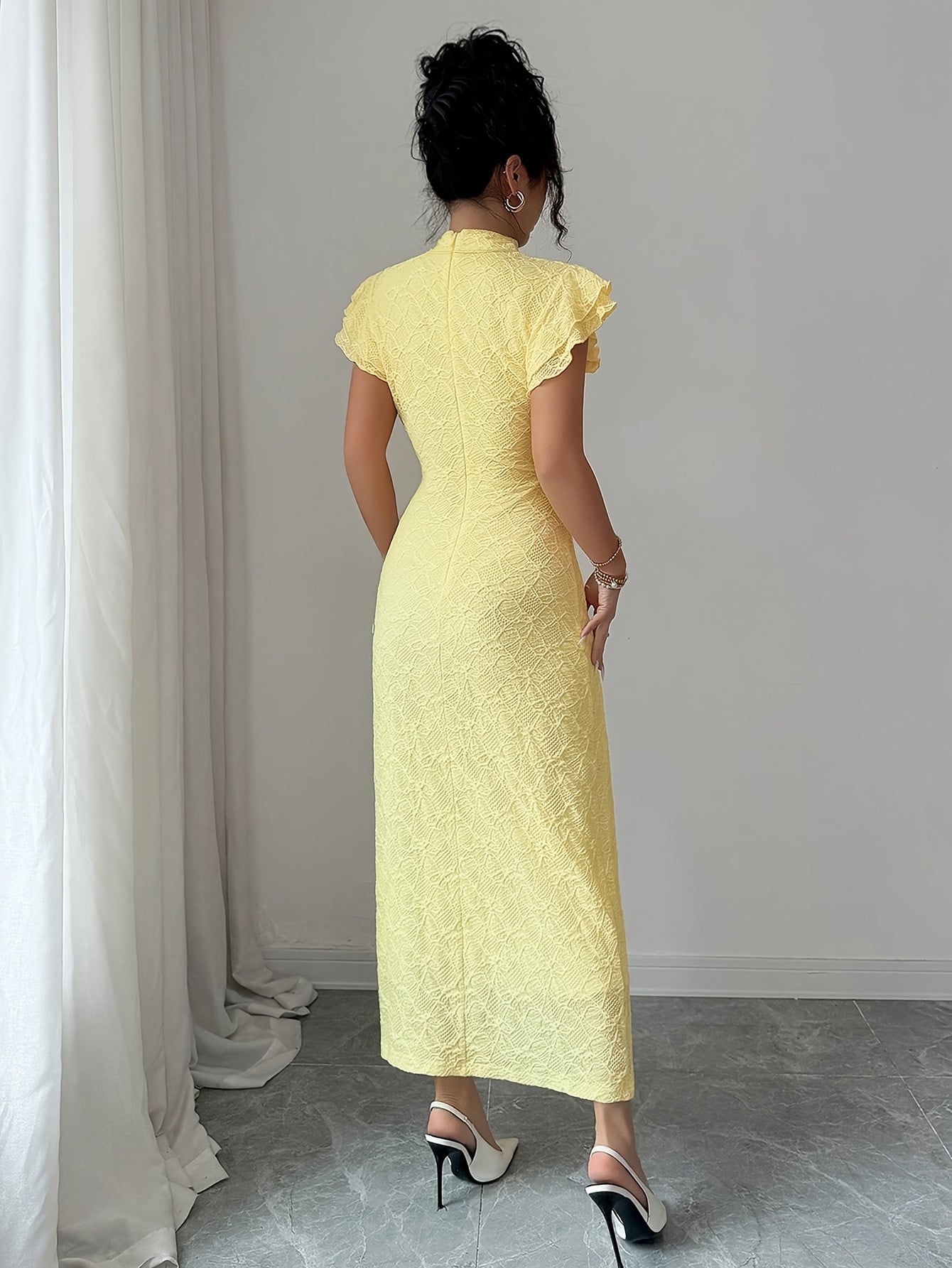 A woman viewed from behind wearing a pale yellow textured cheongsam-style dress with short flutter sleeves and high heels in a minimalist indoor setting.