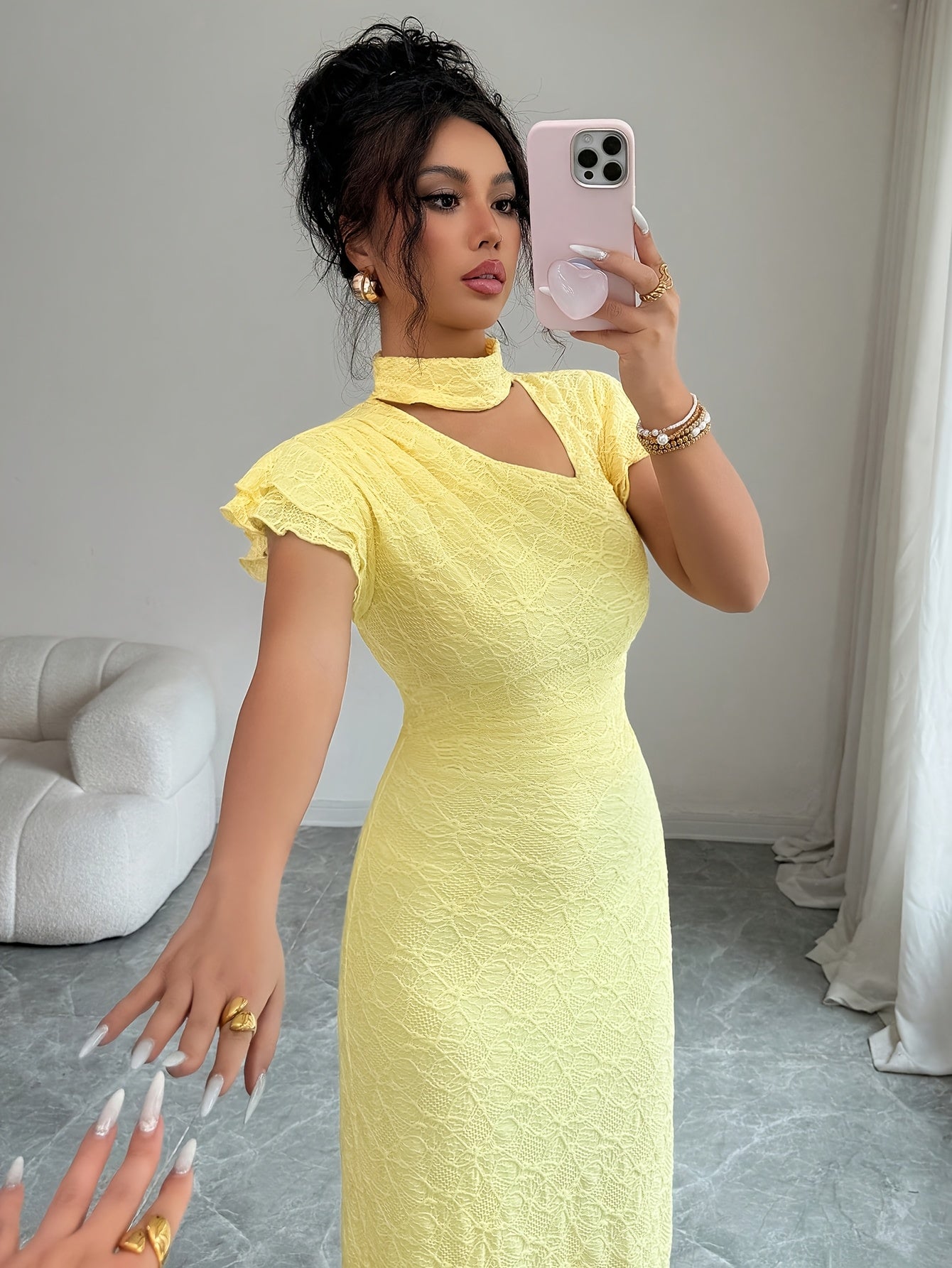 A woman takes a mirror selfie wearing a pale yellow textured midi dress with a mock neck, short flutter sleeves, and a side cutout. She is holding a light pink phone in a modern room with gray flooring and a white sofa.