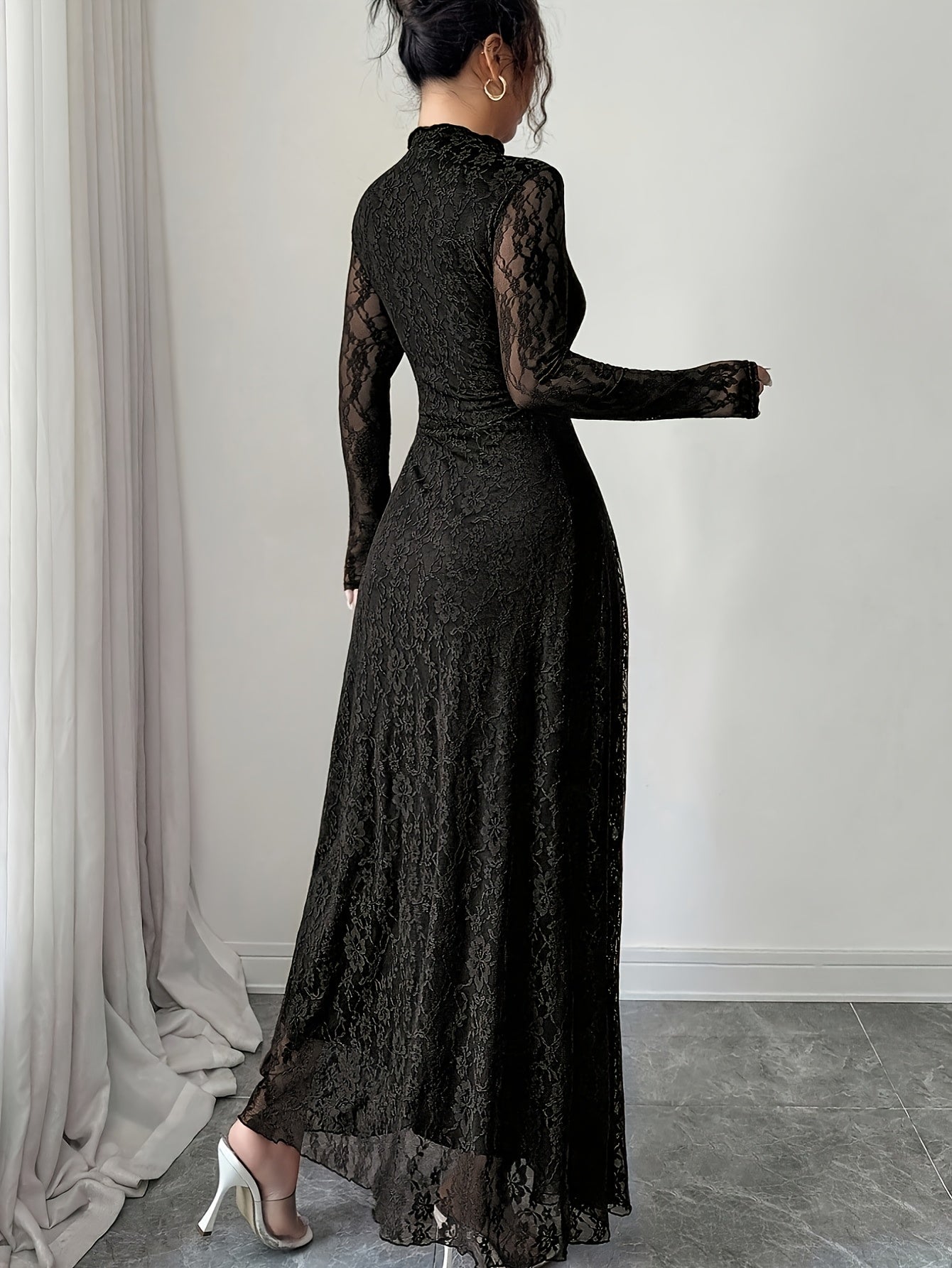 A woman wearing a long-sleeved black lace maxi dress, shown from the back in a minimalist indoor setting with a gray floor and white walls.