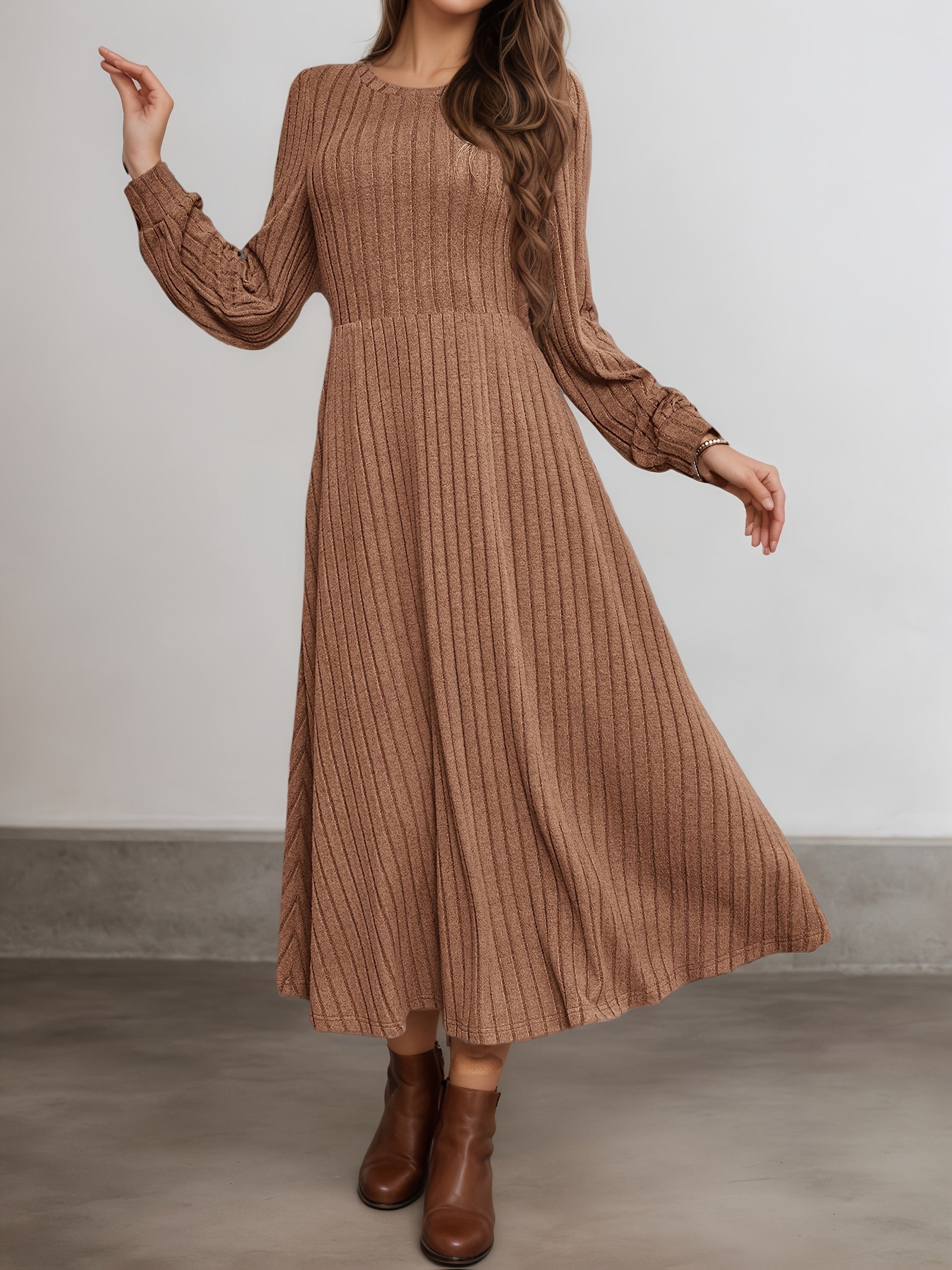 A woman wearing a long-sleeve, midi-length brown ribbed knit dress with a round neckline and flared skirt, paired with brown ankle boots, standing against a neutral gray wall.