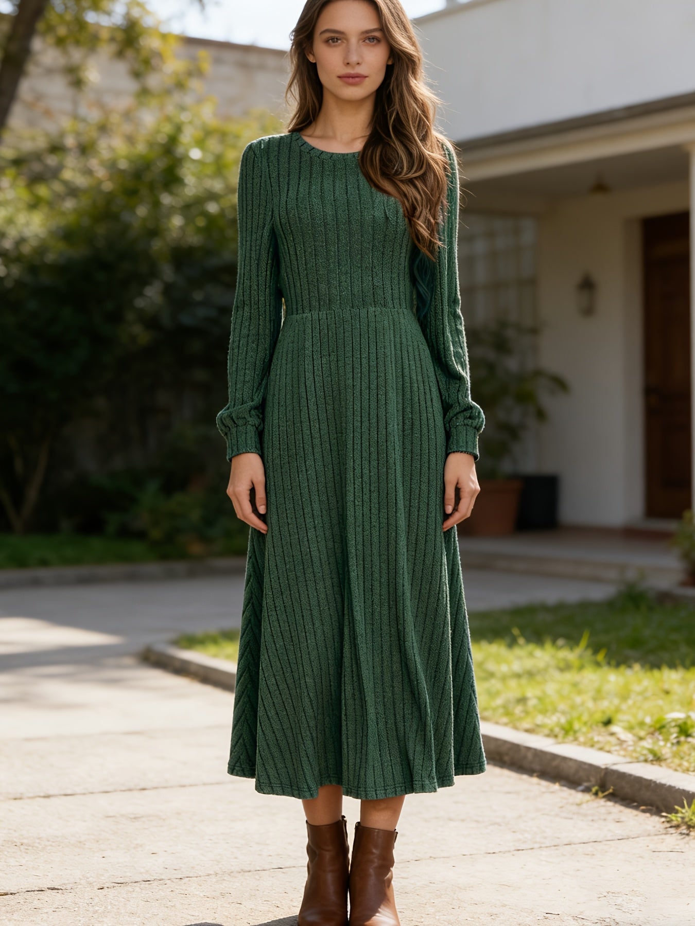 A woman wearing a dark green ribbed knit long-sleeve midi dress with a round neckline and gathered waist, paired with brown ankle boots, standing outdoors on a paved path in front of a house.