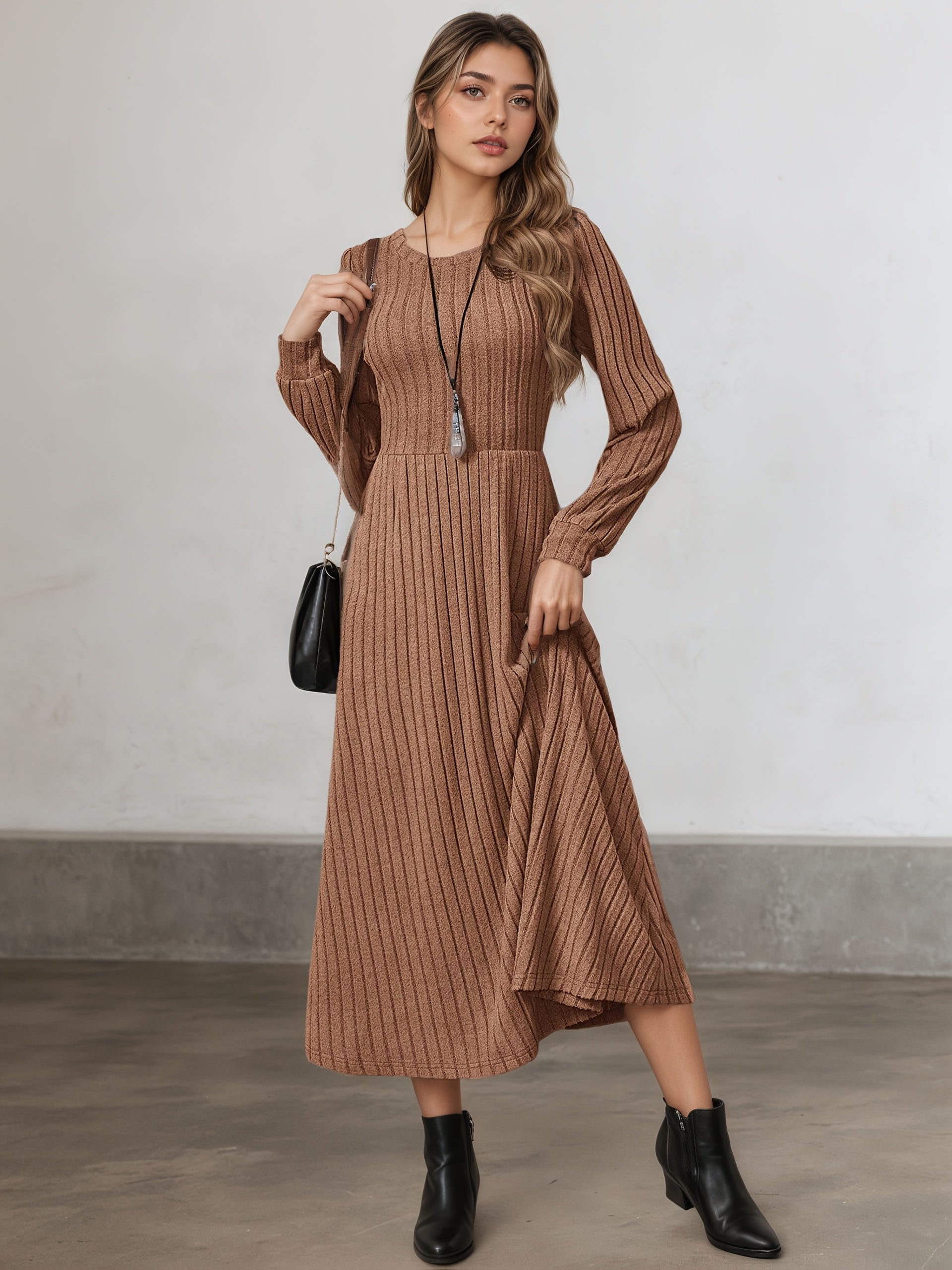 A woman models a long-sleeve, brown ribbed midi dress with a high-low hem, paired with black ankle boots and a small black handbag, standing against a neutral gray wall.