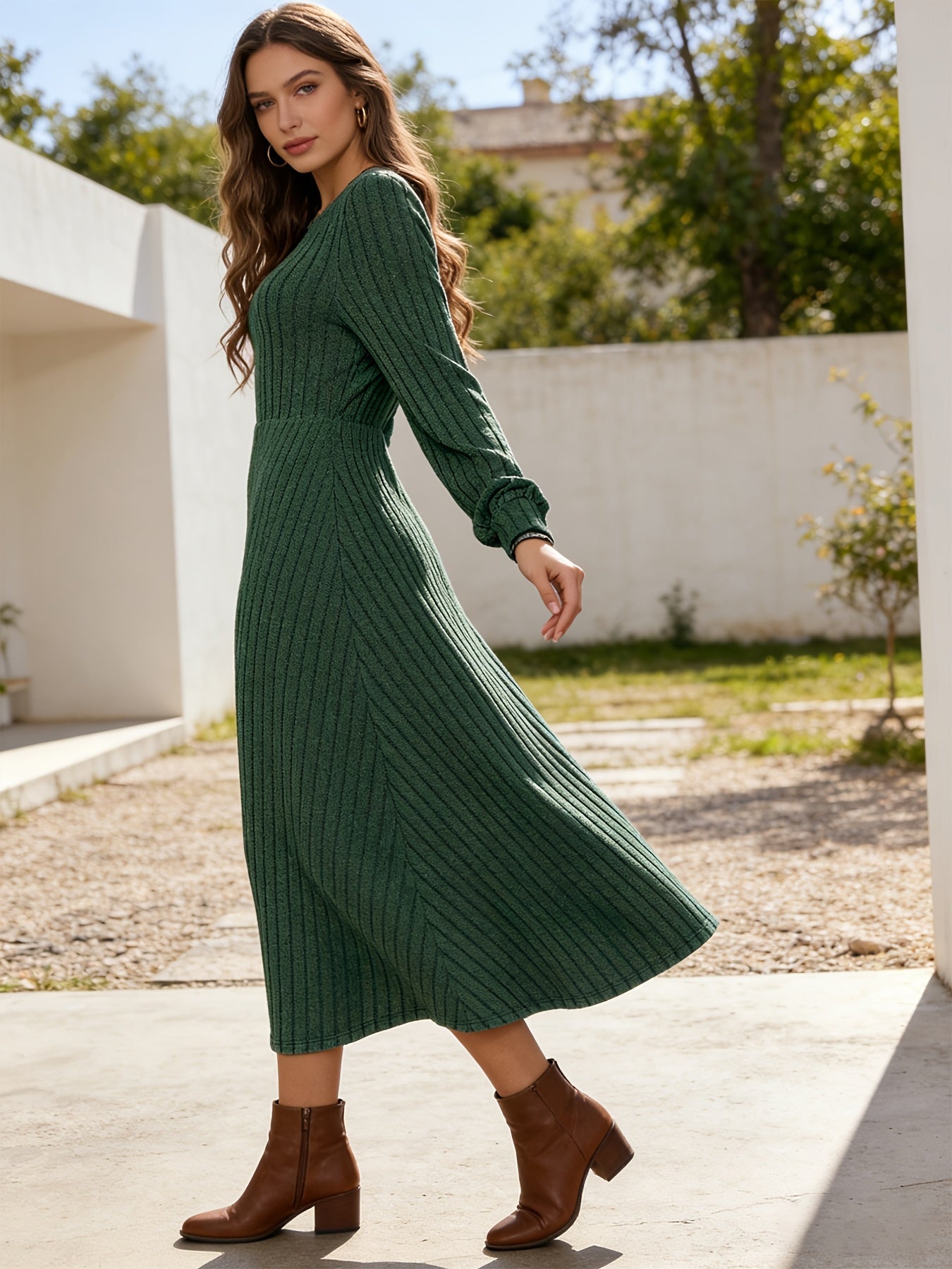 A woman wearing a dark green ribbed knit midi dress with long sleeves and brown ankle boots, standing outdoors in a sunny courtyard with white walls and green trees in the background.