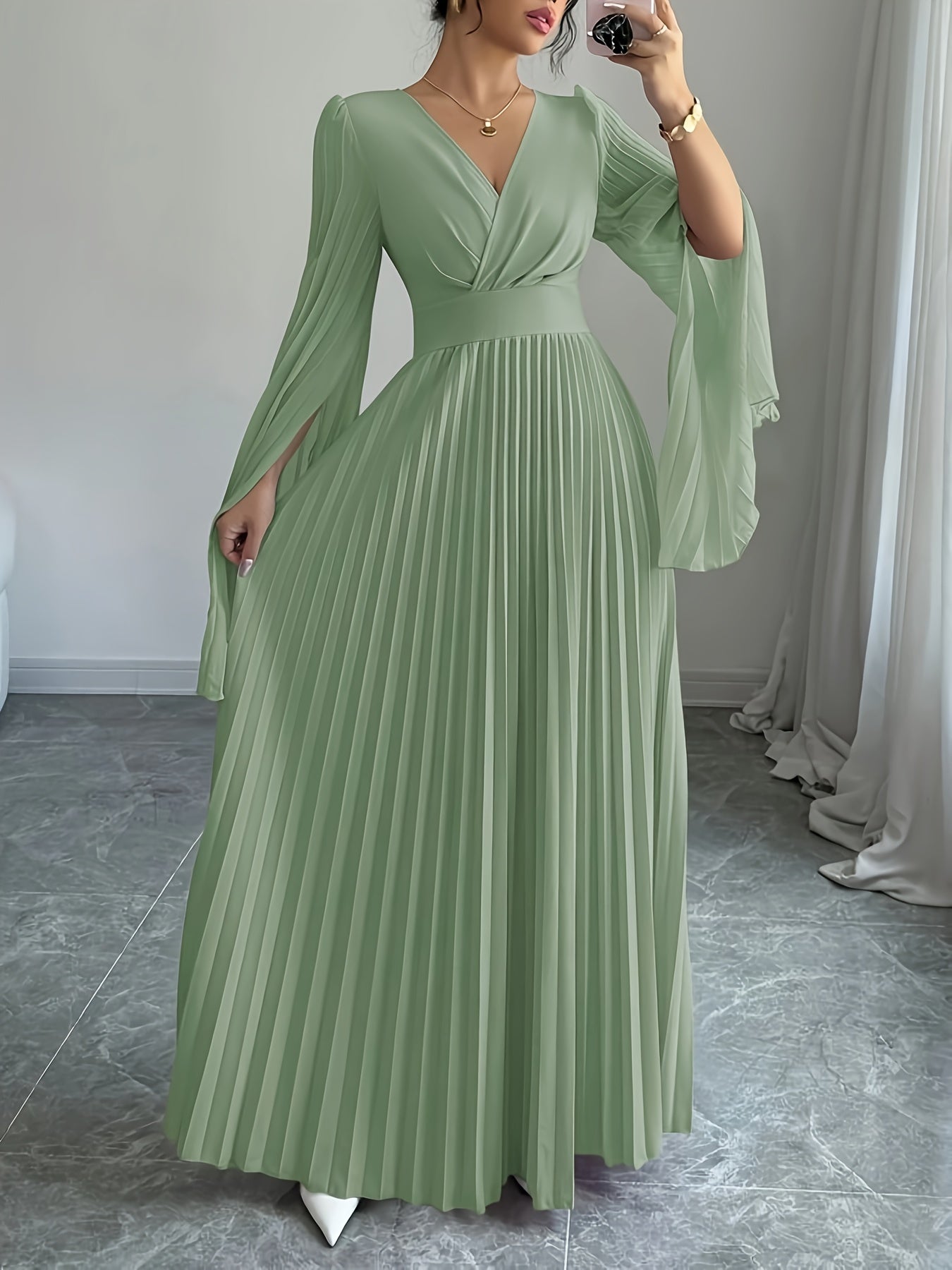 A woman wearing a sage green maxi dress with a pleated skirt, V-neckline, and flared sleeves, taking a mirror selfie in an indoor setting.