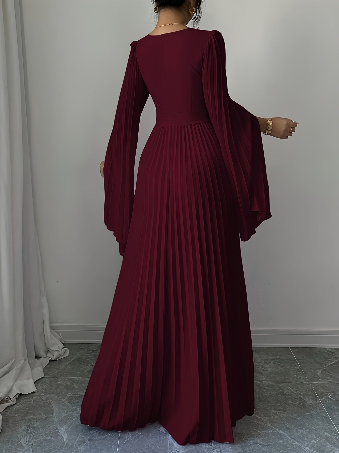 A woman wearing a floor-length burgundy maxi dress with long draped sleeves and a pleated skirt, shown from the back in an indoor setting with a light gray wall and tiled floor.