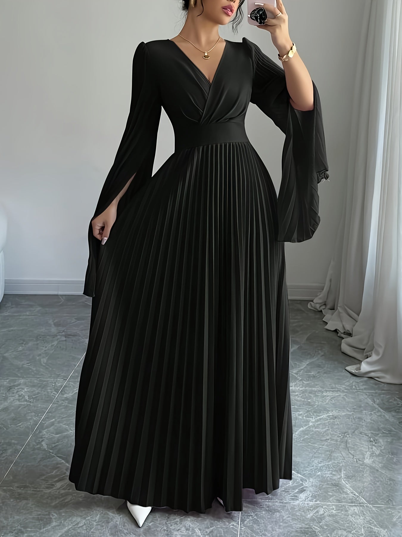 A woman wearing a long black pleated maxi dress with a V-neckline, wide draped sleeves, and a fitted waist, photographed indoors against a light gray wall and floor.