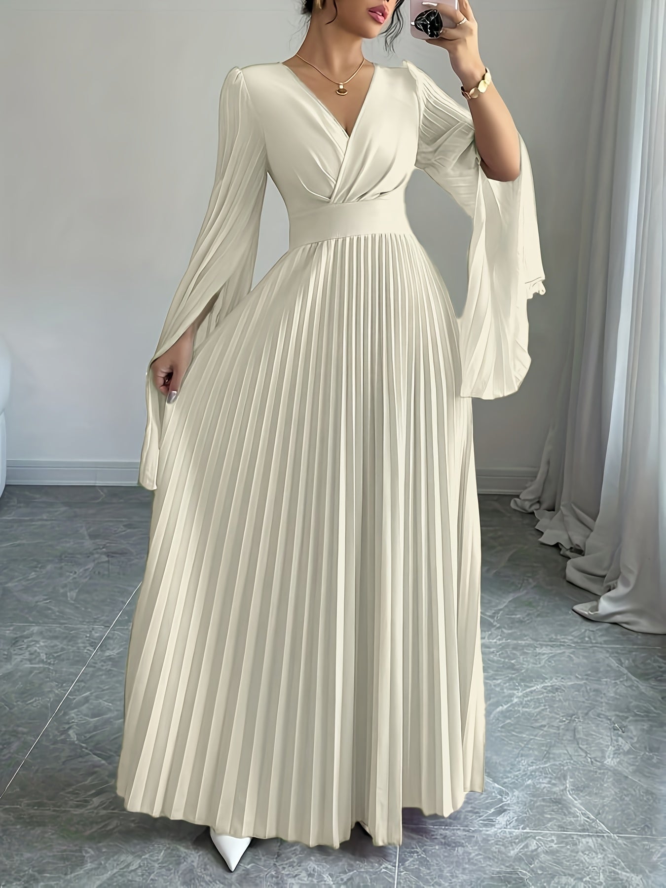 A woman wearing a floor-length cream pleated dress with a V-neckline, fitted waist, and dramatic flared sleeves, photographed indoors against a light gray wall and curtain.