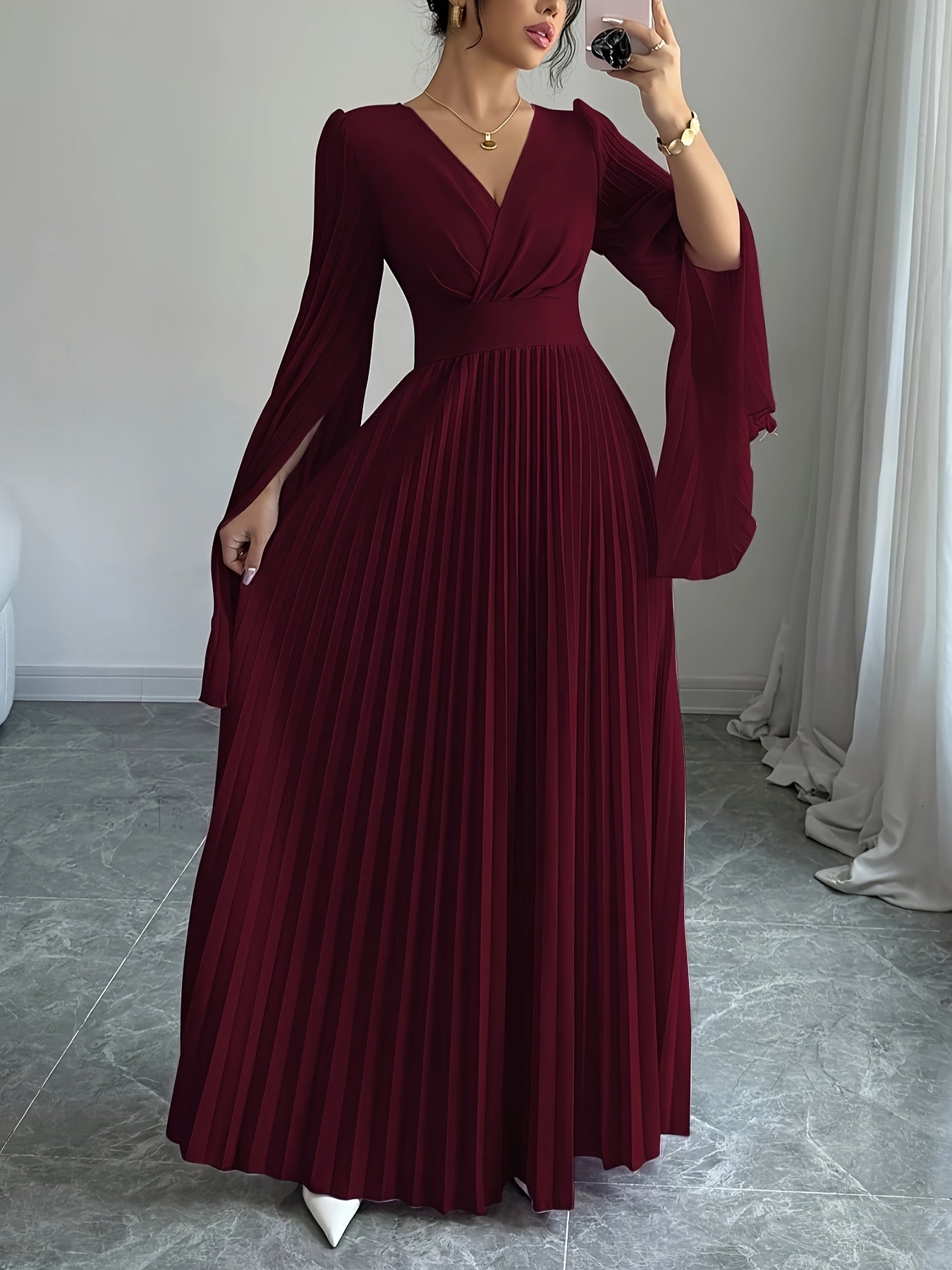 A woman takes a mirror selfie wearing a long burgundy maxi dress with a V-neckline, draped sleeves, and a pleated skirt, standing in a room with gray tiled floors and white curtains.