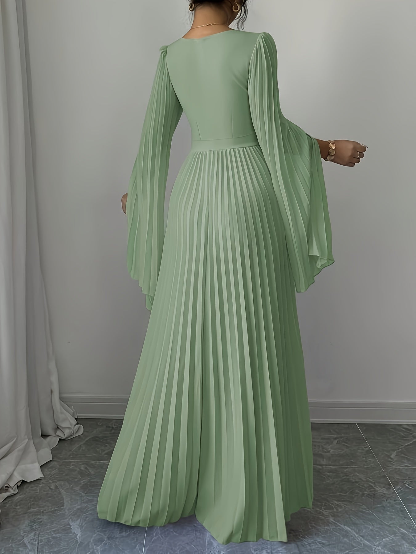 A woman wearing a sage green maxi dress with long flowing sleeves and a pleated skirt, shown from the back in an indoor setting with a gray wall and tiled floor.