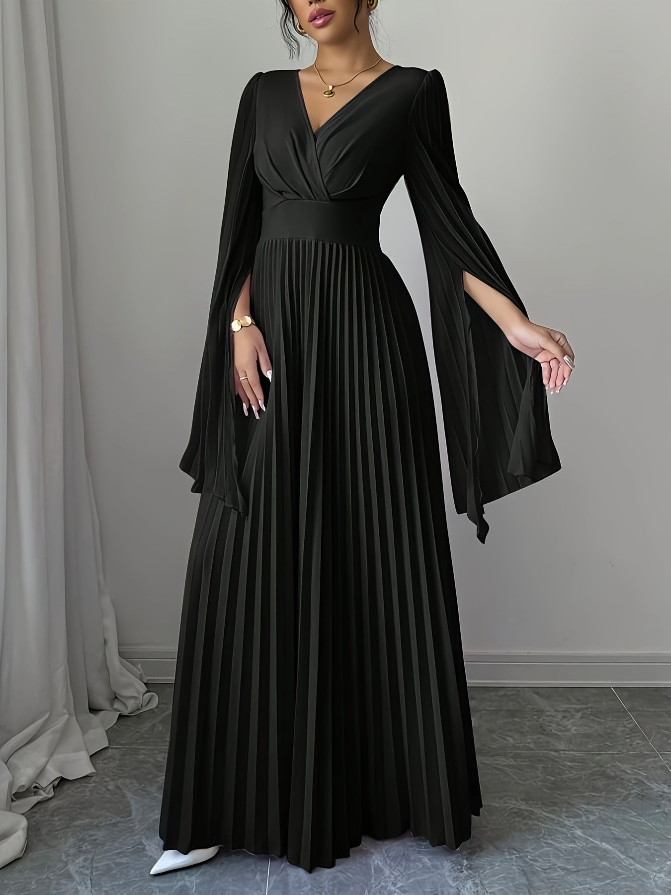 A woman wearing a long black pleated dress with a wrap-style V-neck bodice, sheer draped sleeves, and a fitted waist, standing in an indoor room with gray walls and a gray floor.