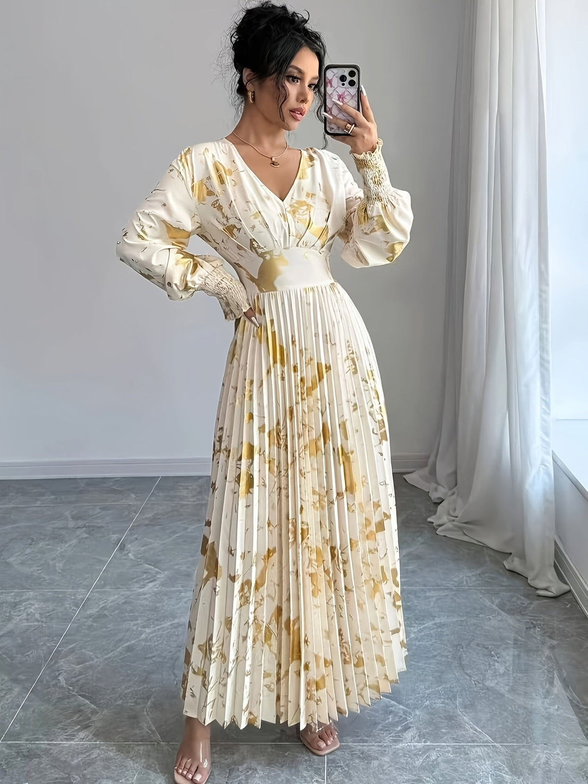 Woman taking a mirror selfie wearing a long floral dress in a room with light gray walls and a white curtain.