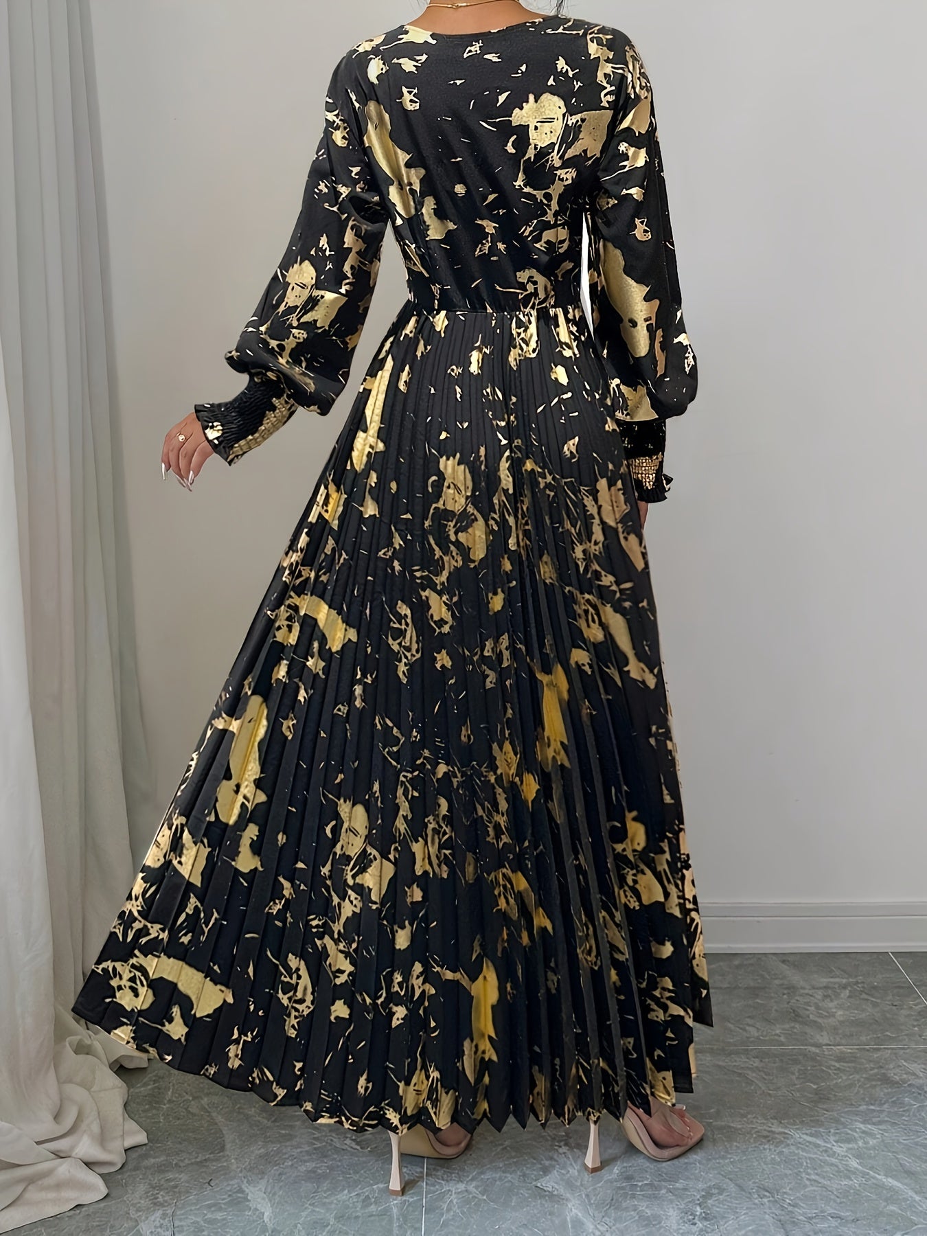 A woman modeling a long black maxi dress with a gold abstract floral print, shown from the back in an indoor setting with a gray tiled floor and light gray wall.