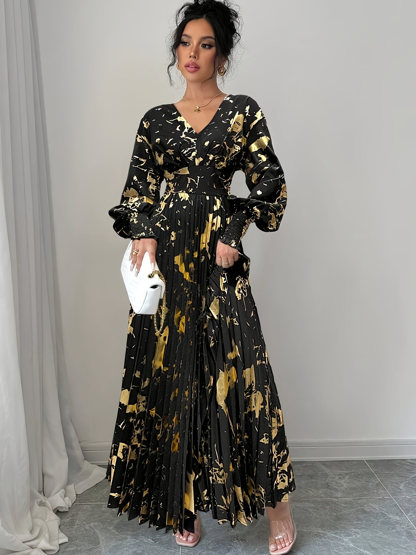 A woman models a long black maxi dress with a gold abstract print, featuring balloon sleeves, a V-neckline, and a pleated skirt. She is holding a white clutch and wearing clear high-heeled sandals, standing against a neutral gray wall.