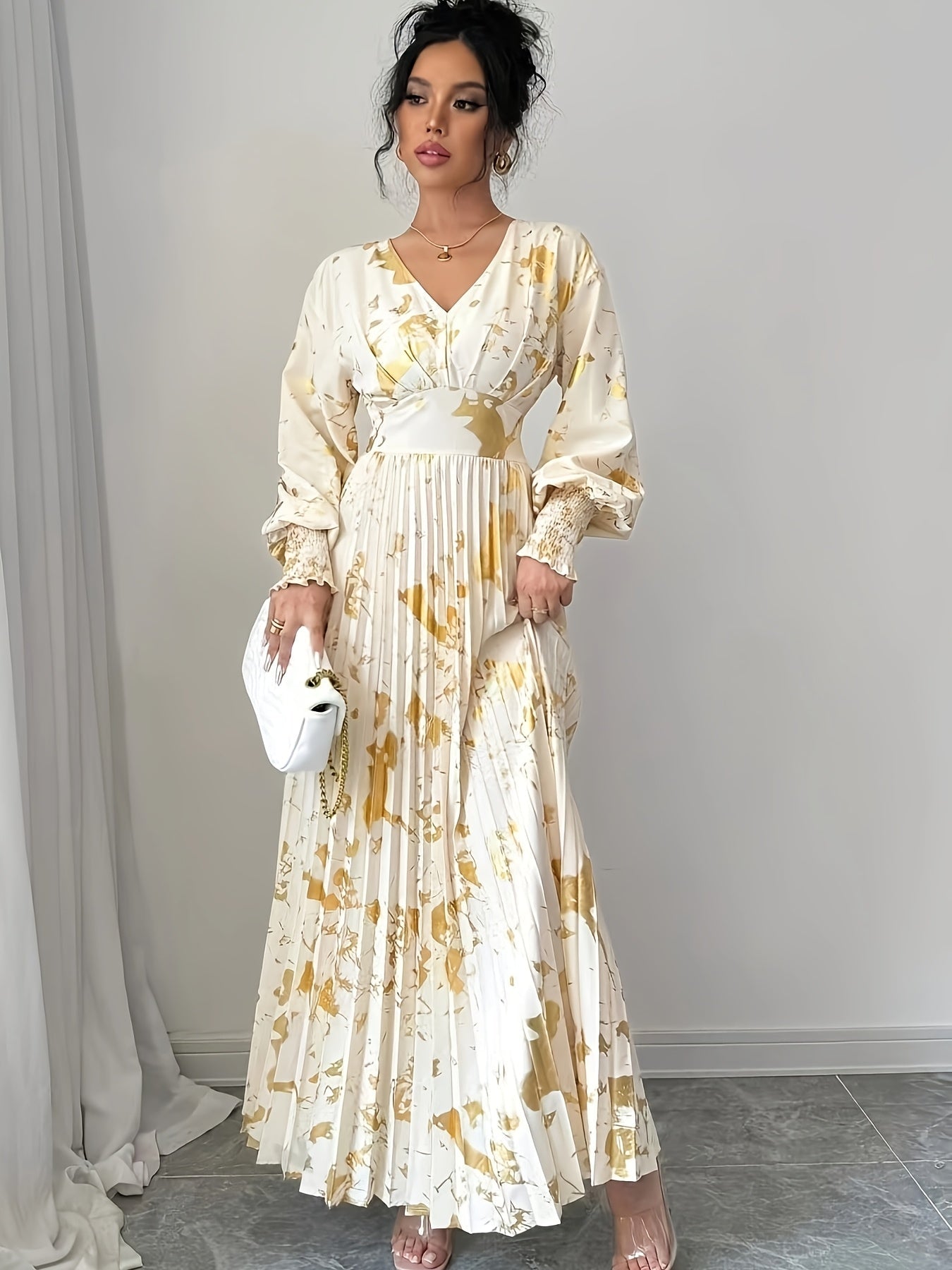 A woman models a long-sleeved cream maxi dress with a gold abstract floral print, featuring a pleated skirt and a gathered waist. She is holding a small white handbag and standing in front of a neutral gray wall.