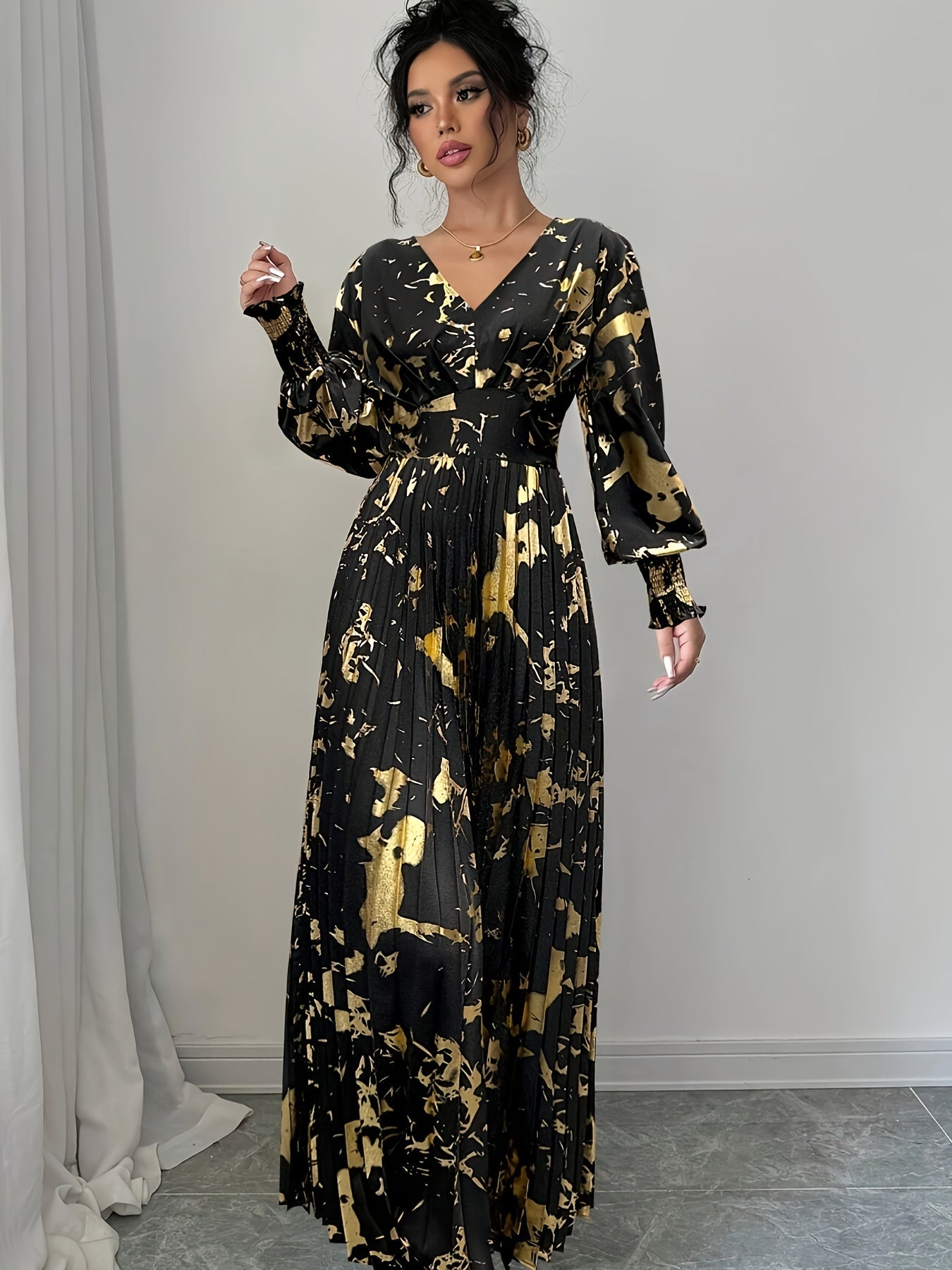 A woman models a long-sleeved black maxi dress with a gold abstract painterly print, featuring a pleated bodice and skirt, set against a neutral indoor background.