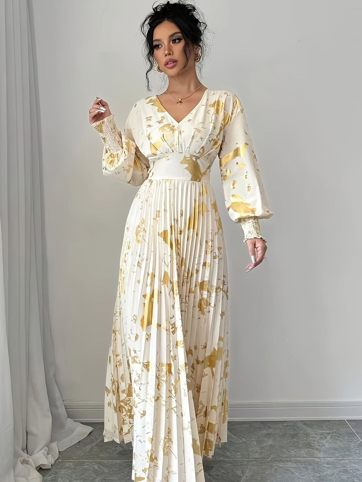 A woman models a long-sleeved cream maxi dress with a yellow floral print and pleated skirt, standing in a minimalist room with a gray floor and white curtain.