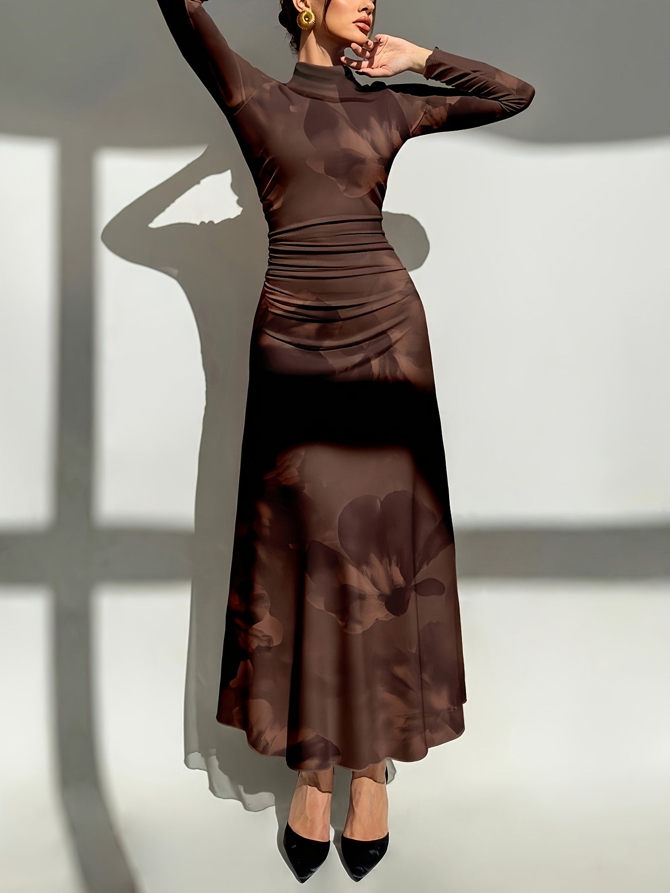 A woman models a long-sleeve, high-neck, floor-length brown dress with a subtle tonal floral pattern and ruched detailing at the waist, posed against a light gray wall with strong window shadows.