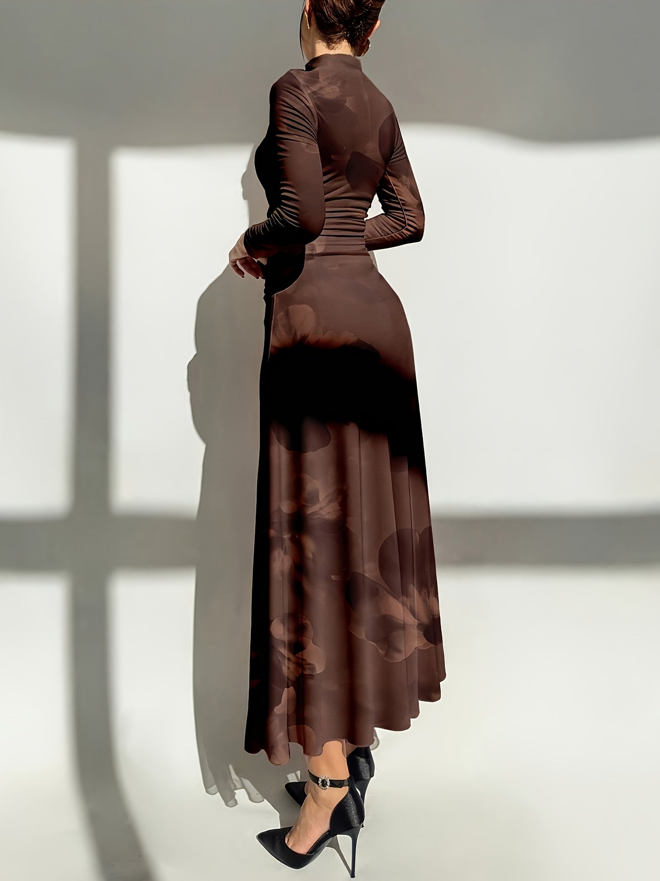 A woman shown from the back wearing a long-sleeve, high-neck, floor-length brown dress with a subtle tonal floral pattern and black high heels, standing against a white wall with strong shadow patterns.