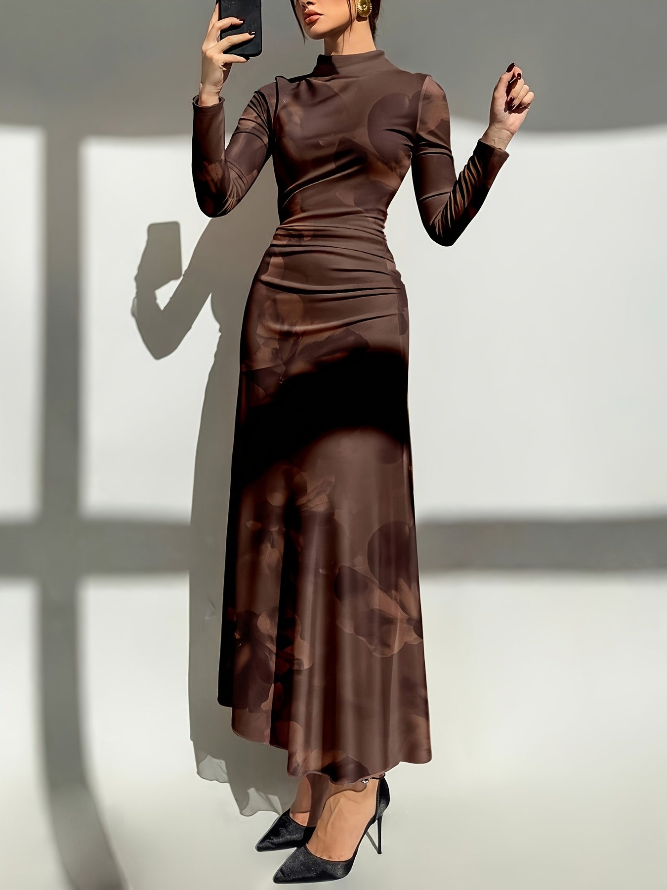 A woman modeling a long-sleeve mock-neck maxi dress in a dark brown color with a subtle tonal abstract pattern, paired with black pointed-toe heels, set against a white wall with strong window shadows.