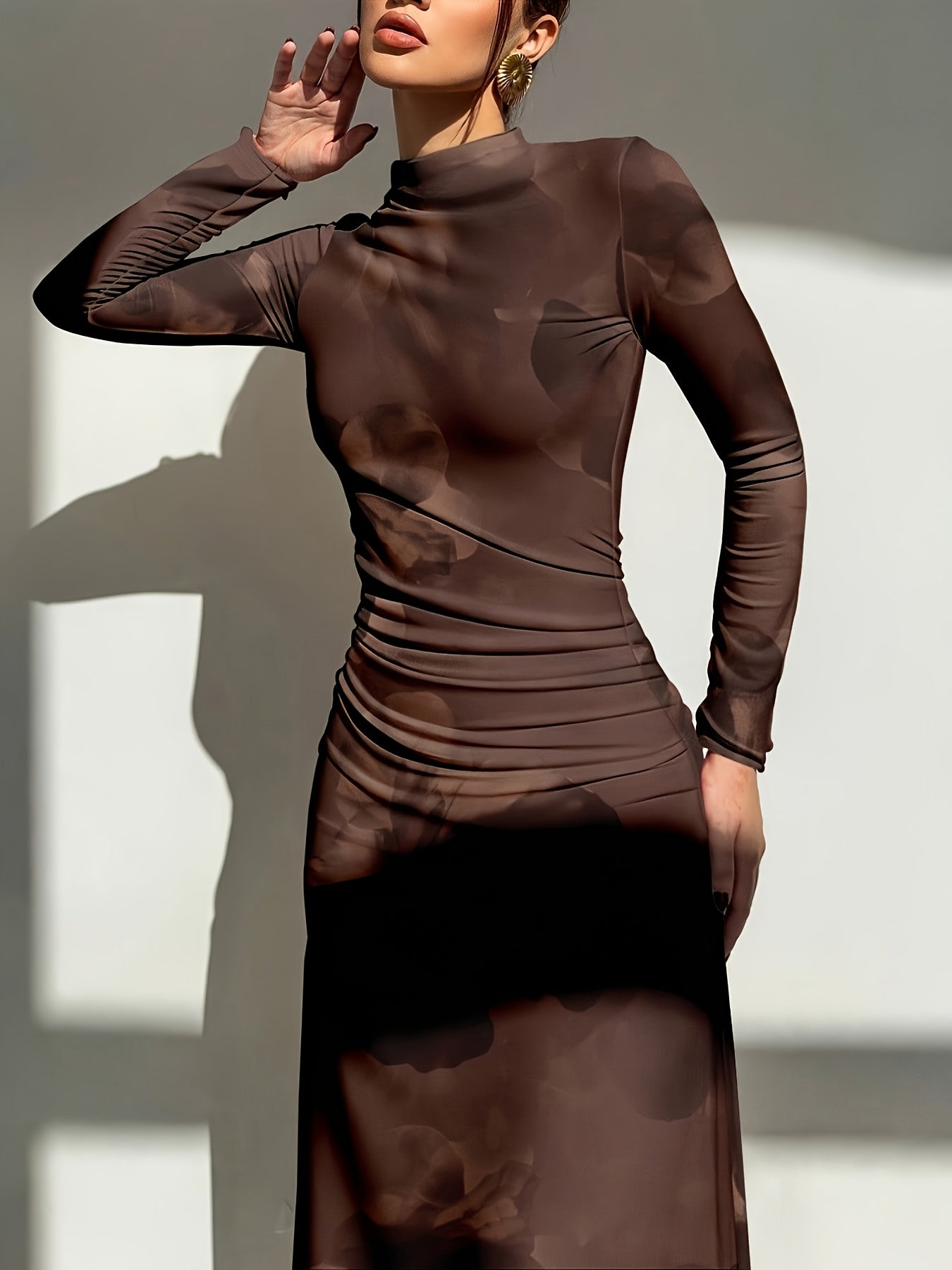 A woman wearing a long-sleeve, high-neck, ruched midi dress in a dark brown abstract camouflage pattern, posed against a wall with dramatic sunlight and shadows.