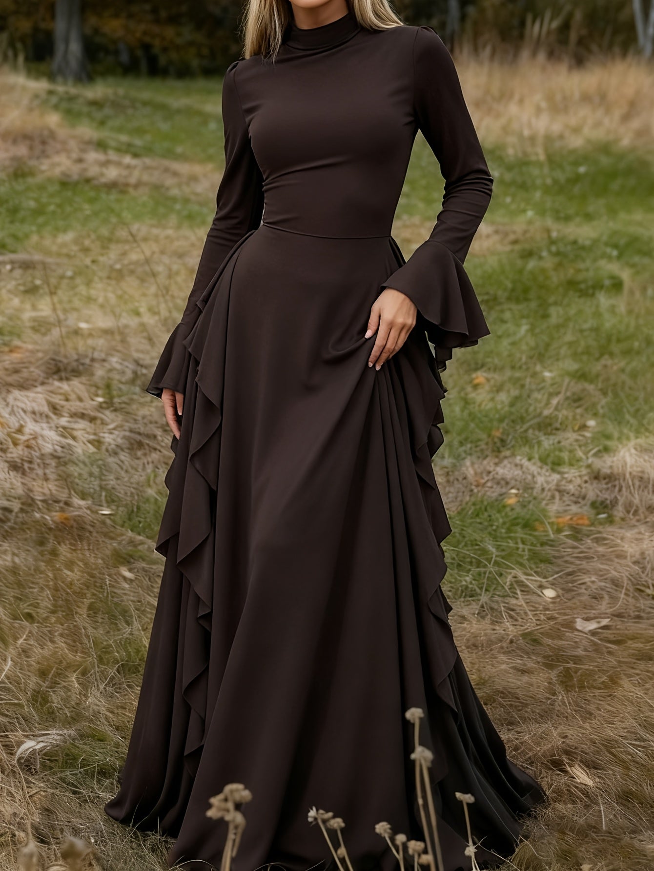 A woman wearing a long-sleeve dark brown high-neck maxi dress with flared cuffs and a tiered ruffle skirt, standing outdoors in a grassy field.