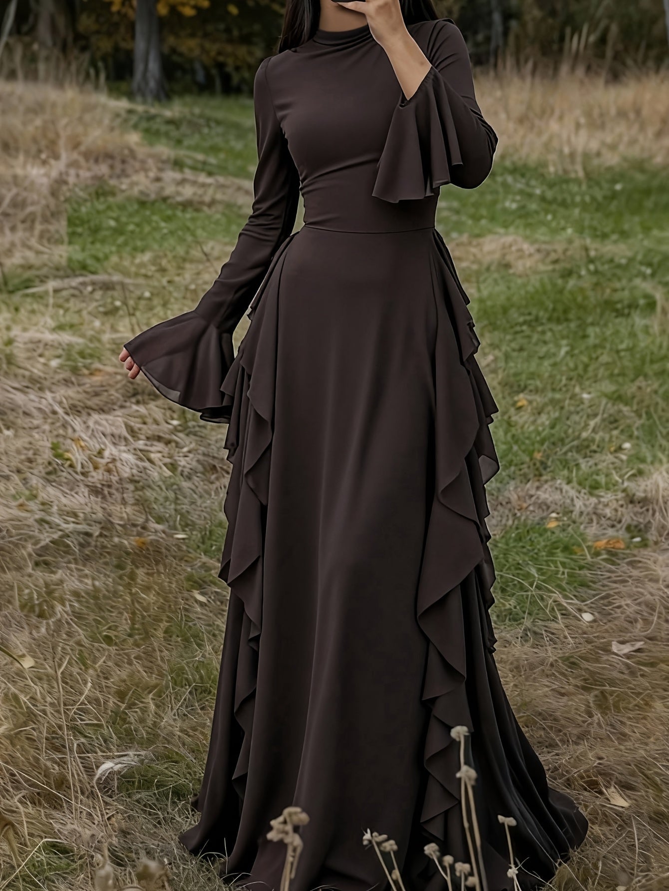 A woman wearing a long-sleeve dark brown maxi dress with dramatic ruffled layers and flared sleeves, standing outdoors in a grassy field.