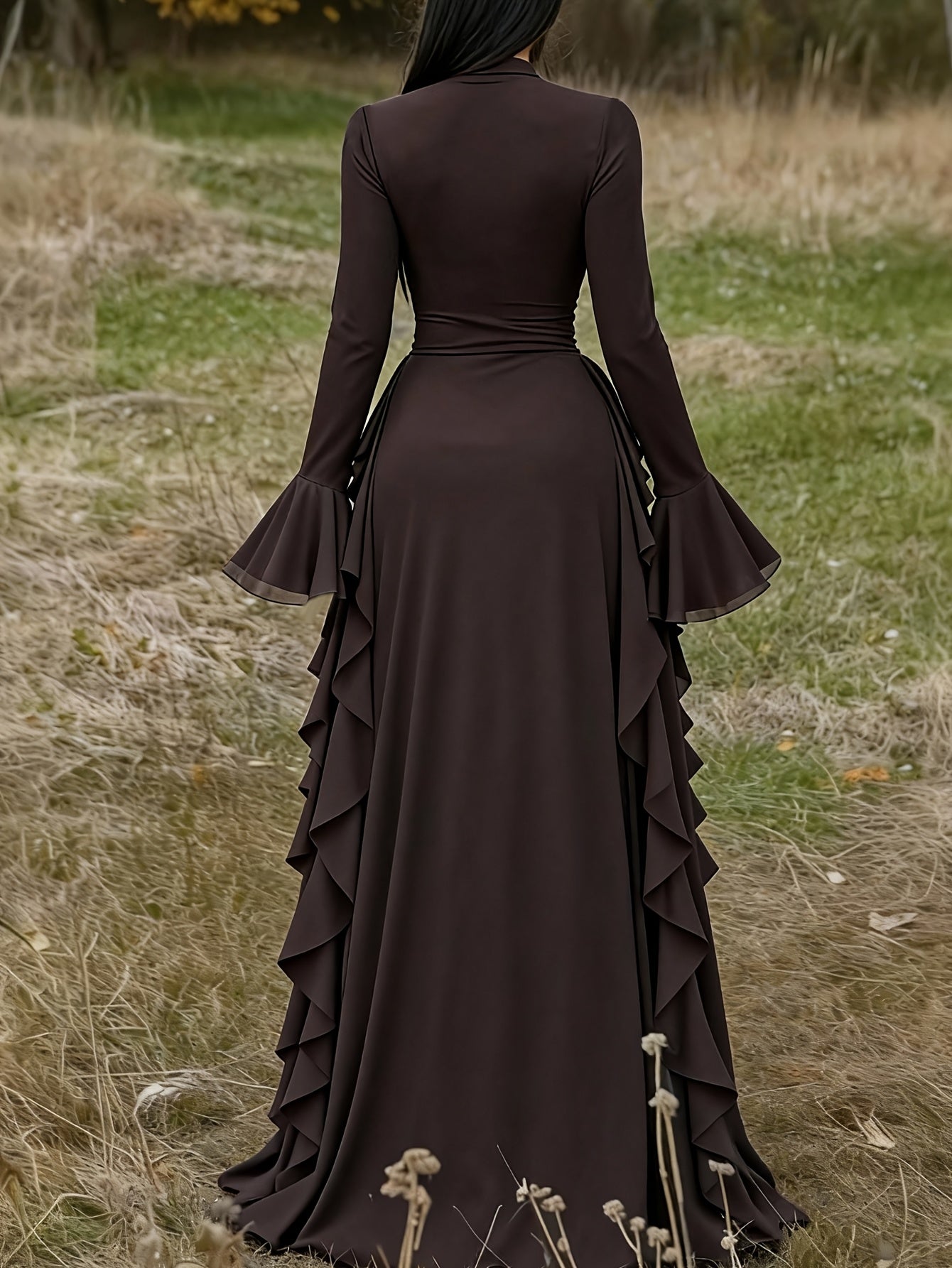 A woman stands outdoors in a grassy field wearing a long-sleeve dark brown maxi dress with flared bell sleeves and layered ruffle details along the sides of the skirt.