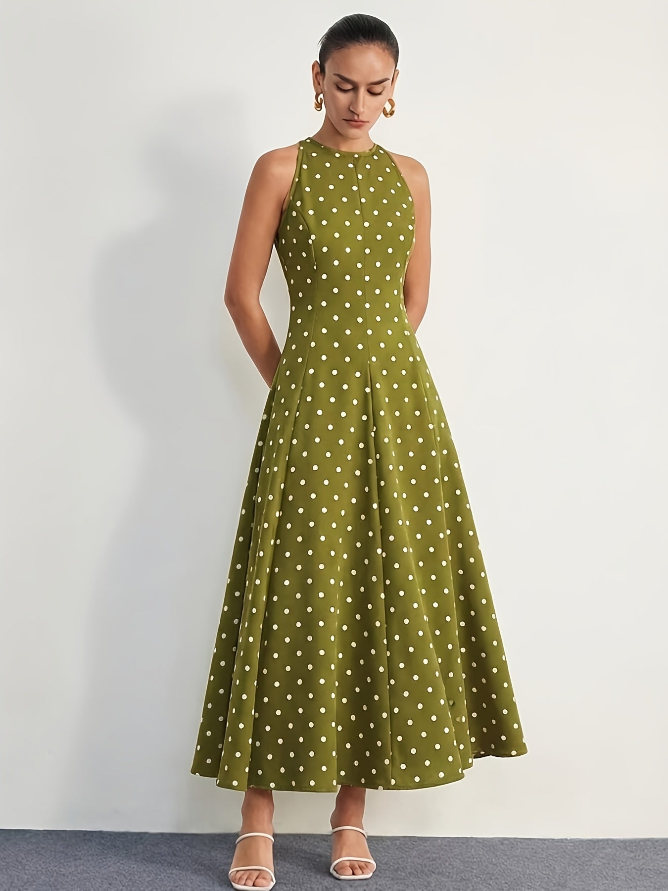 A woman modeling a sleeveless olive green halter maxi dress with a white polka dot pattern, standing against a plain light gray wall.