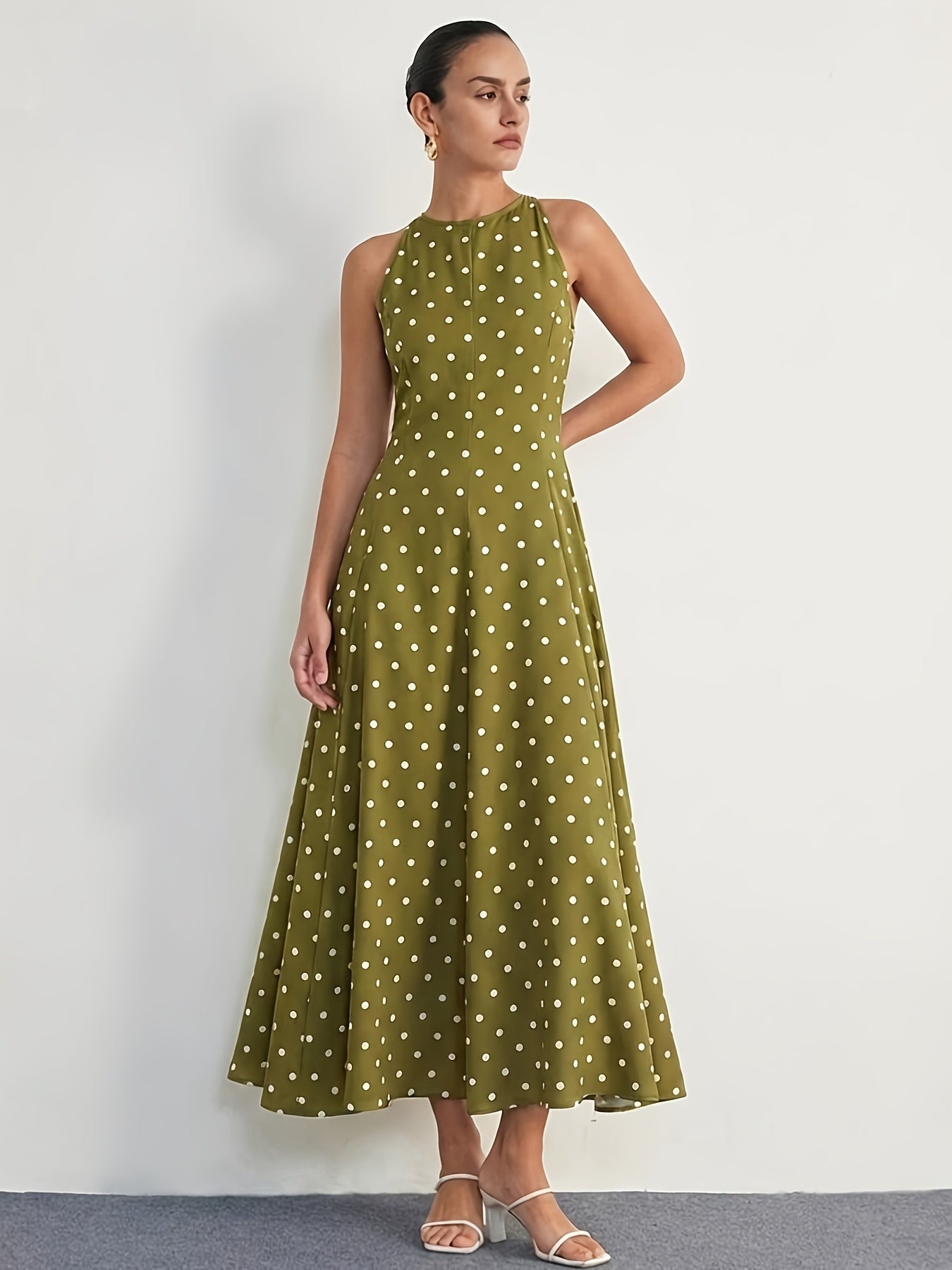 A woman modeling a sleeveless olive green halter-neck maxi dress with a white polka dot pattern, standing against a plain white wall.