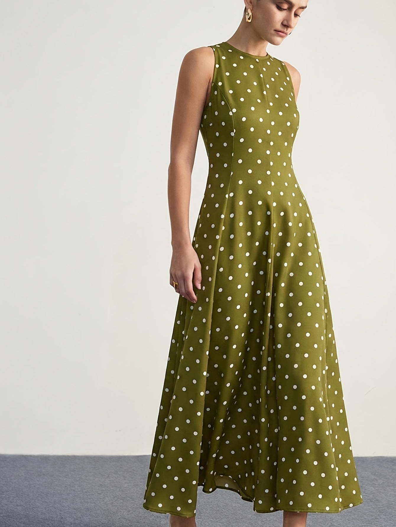 A woman wearing a sleeveless olive green midi dress with a white polka dot pattern, photographed against a plain off-white studio background.