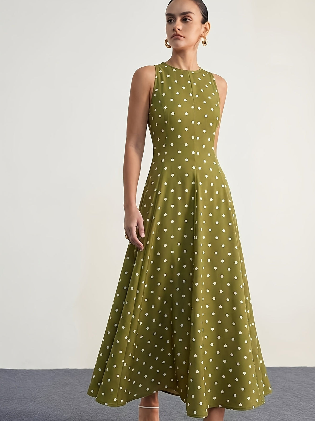 A woman modeling a sleeveless olive green midi dress with a white polka dot pattern against a plain off-white studio background.