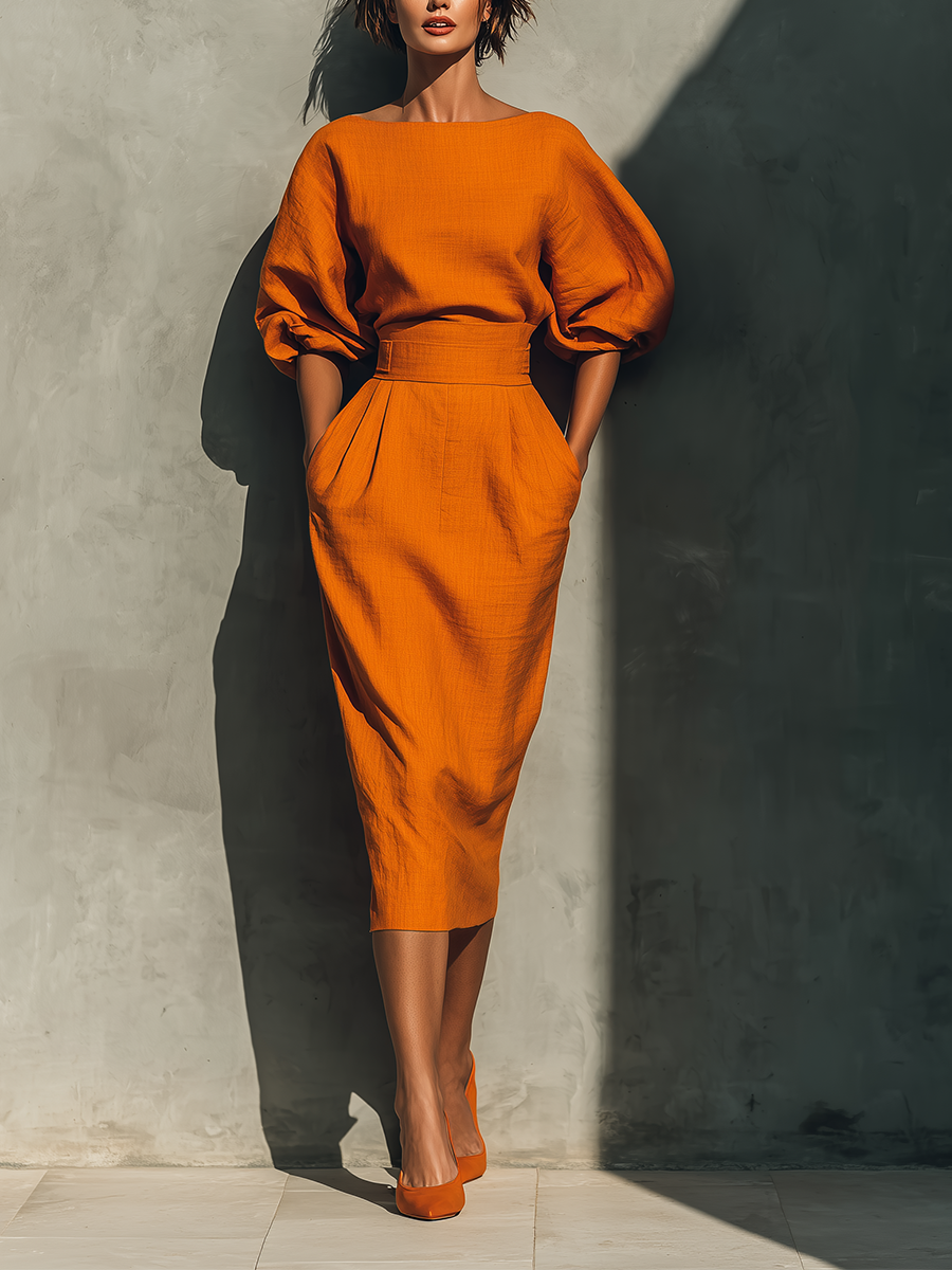 A woman models a bright orange midi dress with voluminous puff sleeves and a matching belt, standing against a textured gray wall with dramatic sunlight and shadows.