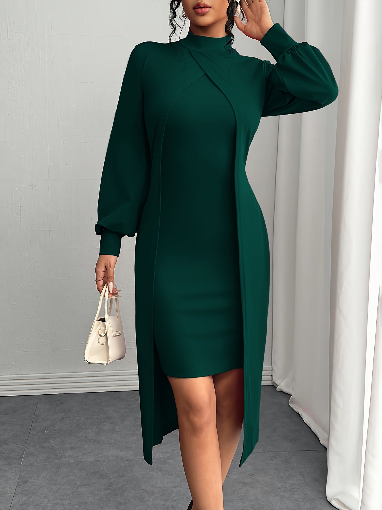 A woman models a dark green long-sleeve midi dress with a high-low front overlay, holding a small cream-colored handbag in a bright indoor setting.