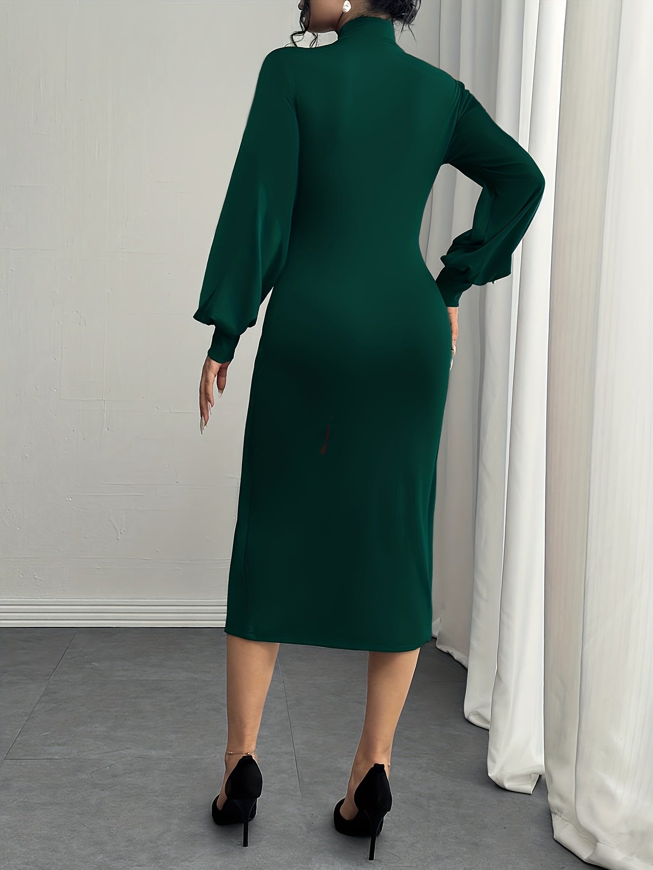 A woman wearing a dark green long-sleeve midi dress, shown from the back. She is standing in a room with a gray floor, a white wall, and white curtains, wearing black high heels.